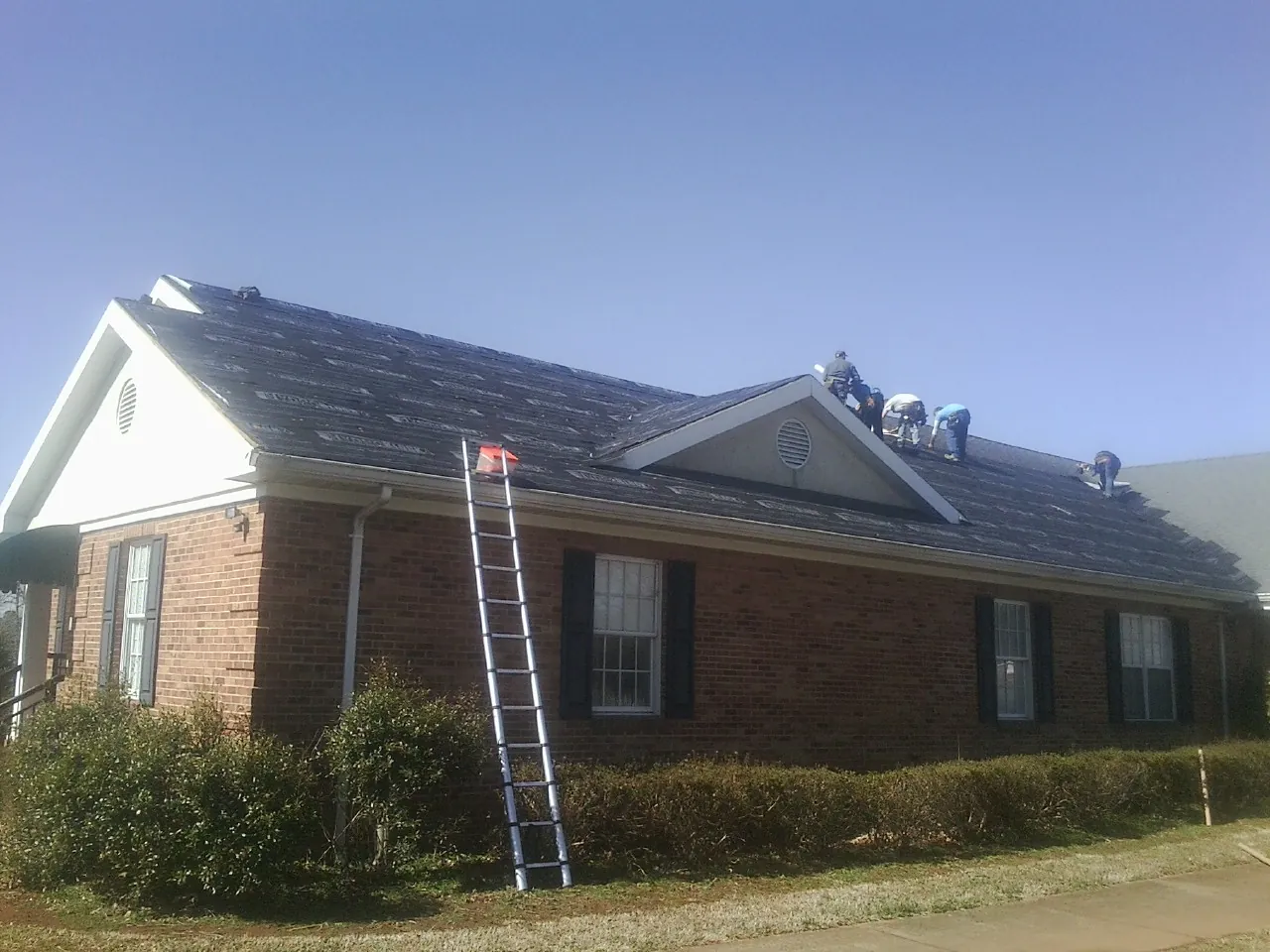 Crew tearing shingles off a brick ranch roof with a ladder set — Emergency Roof Repair work by our crew in Livingston Manor