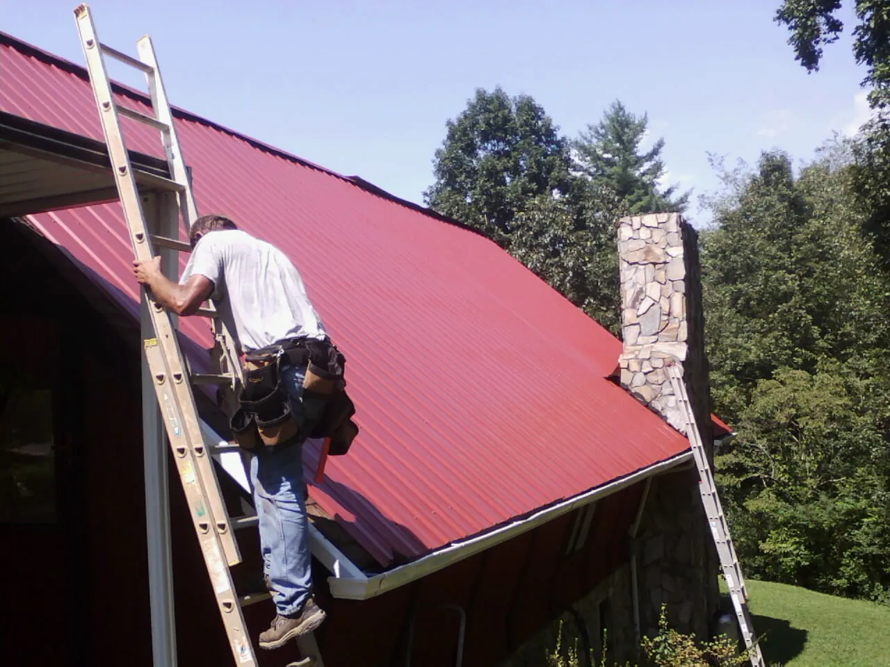 Roofer on a ladder beside a red metal roof and stone chimney — Soffit & Fascia Repair by Roofing Brothers in Livingston Manor